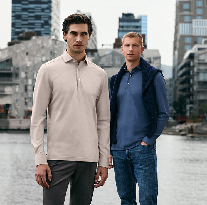 Model in dunkelblauem Jersey-Hemd mit Knopfleiste, kombiniert mit hellblauer Jeans; im Vordergrund unscharf ein zweiter Mann in hellgrünem Hemd.