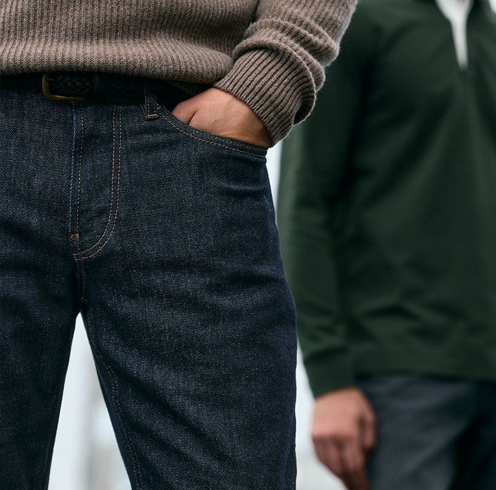 Model in einem weißen T-Shirt, hellblauen Jeans und grauen Schuhen vor einem beigen Hintergrund.