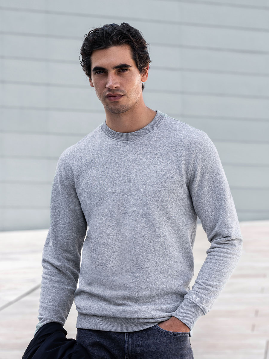 Model in einem grauen Pullover und Jeans, die Arme hinter dem Rücken, leicht vom Objektiv wegschauend. Foto aufgenommen vor einem hellgrauen Hintergrund.