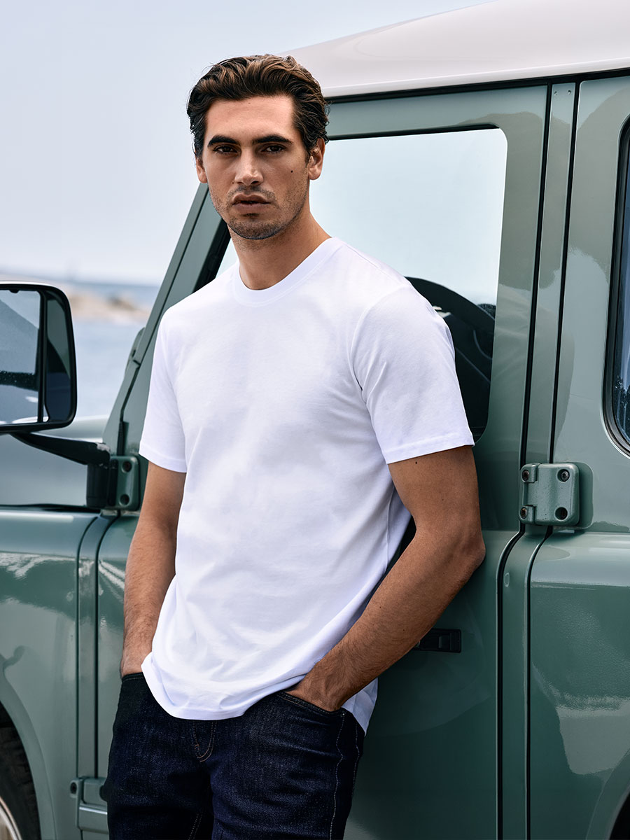 Model in einem weißen T-Shirt mit Rundhalsausschnitt und grauen Jeans, die Arme auf den Beinen abgestützt und direkt in die Kamera blickend. Foto aufgenommen vor einem hellgrauen Hintergrund.