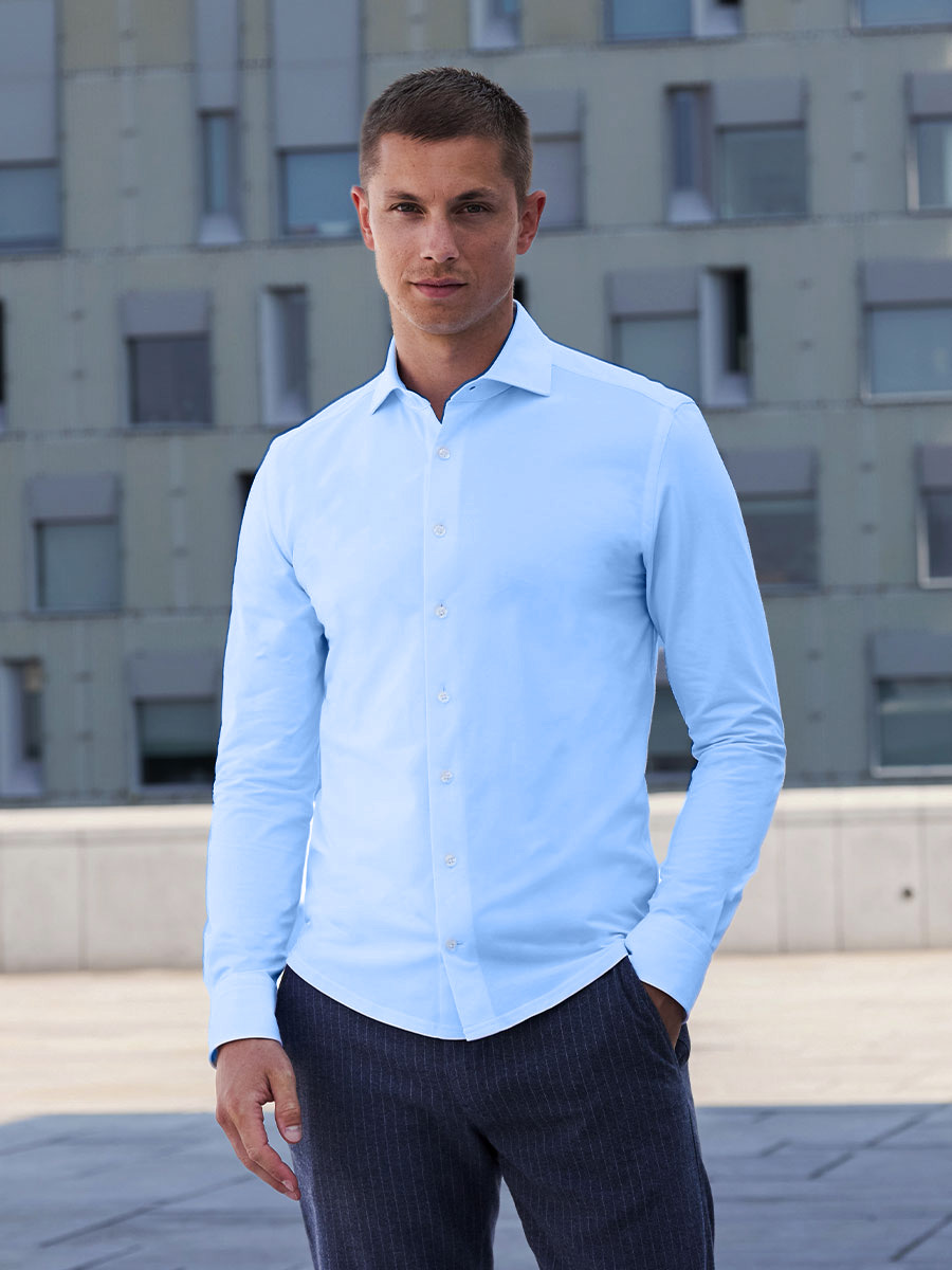 Model in einem weißen Poloshirt und einer blauen Chino, eine Hand über der anderen, direkt in die Kamera blickend. Foto aufgenommen vor einem hellgrauen Hintergrund.
